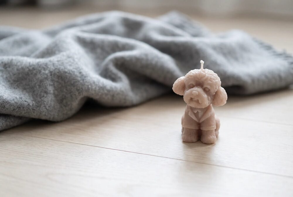 Uszkár alakú bézs gyertya világos parkettán, mögötte szürke gyapjútakaró