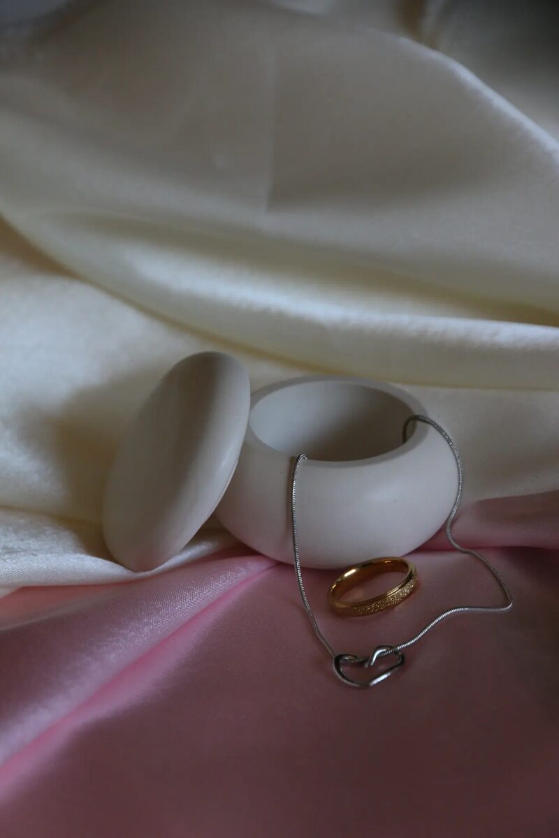 Kavics formájú fehér kerámia tégely levett fedéllel, krém és rózsaszín szaténon.
