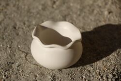 Fehér, gömbölyded kerámia üst hullámos peremmel, napfényben, kavicsos felületen.